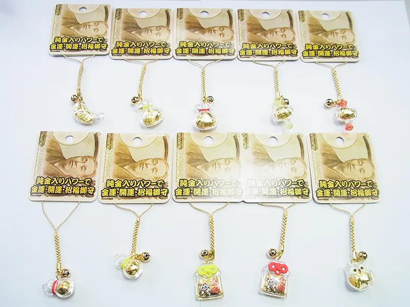 金色の台紙に、鎖と鈴の付いたお守りストラップを上下2段で10点並べたもの。飾りは三日月、巾着袋、小瓶、招き猫入りの小袋など。台紙には「純金入りパワーで 金運・開運・招福御守」の表示