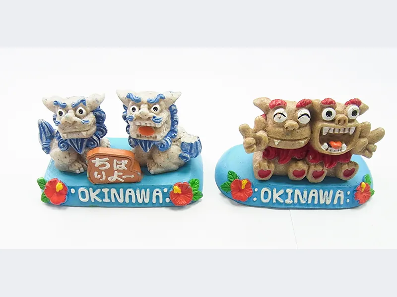 「OKINAWA」と書かれた台座に乗ったシーサーの置物2組。左は青いたてがみの2体と「ちばりよー」の看板、右は赤いたてがみの2体